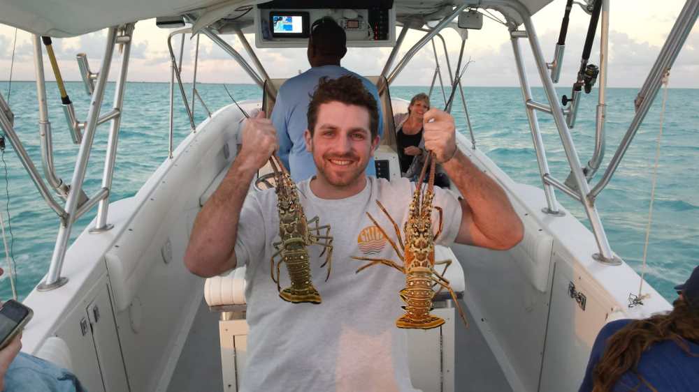 Man on a boat holding two lobsters, with the ocean in the background.